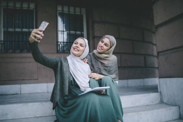 Two women in hijabs take a selfie while sitting on steps, representing life and work opportunities in Dubai.